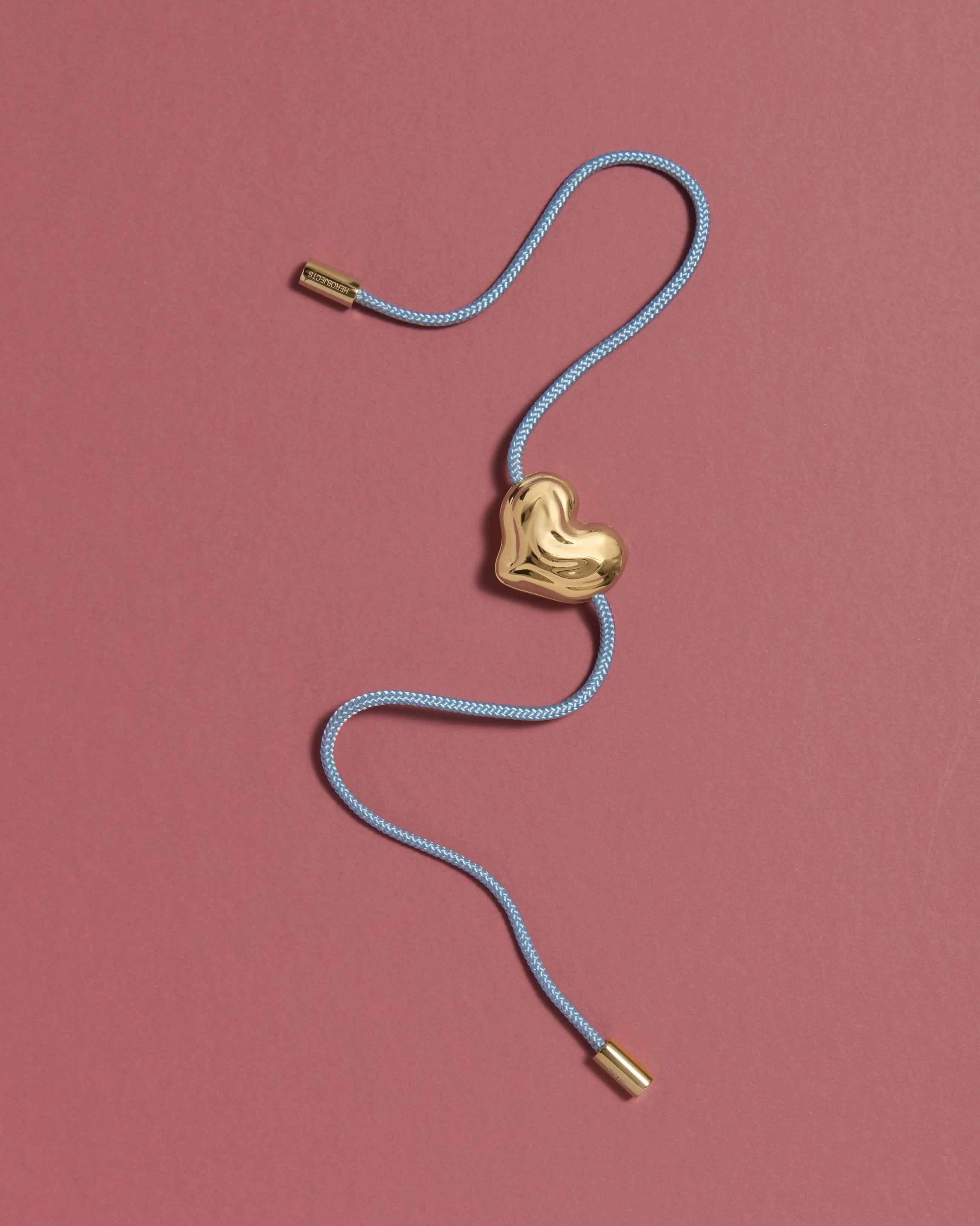 Blue cord necklace with a 18K gold clasp and a heart-shaped 18K gold pendant on a pink background