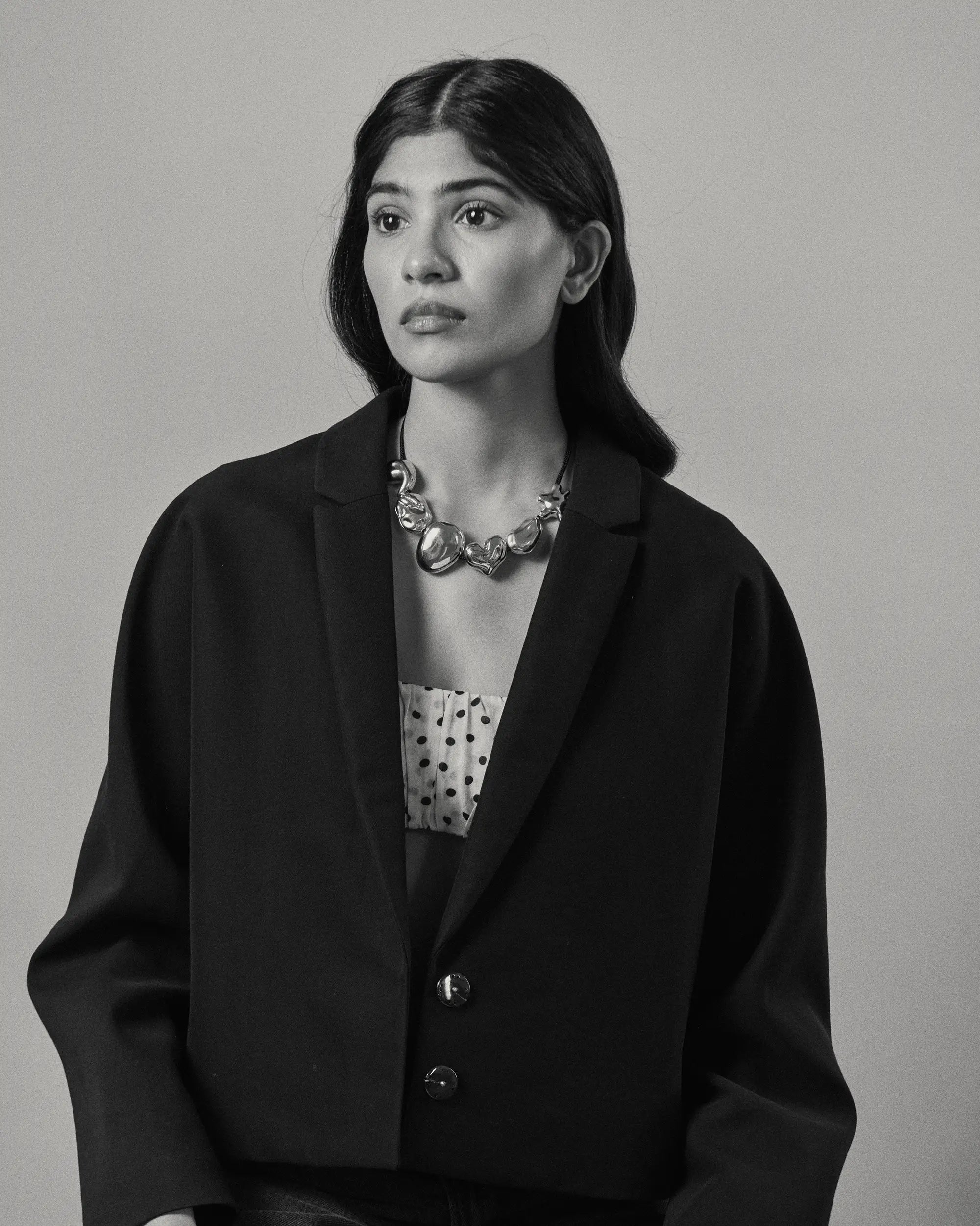 A black and white portrait of a model wearing a black cropped blazer over a white top with black polka dots, accessorized with all 18K Gold Hero objects.