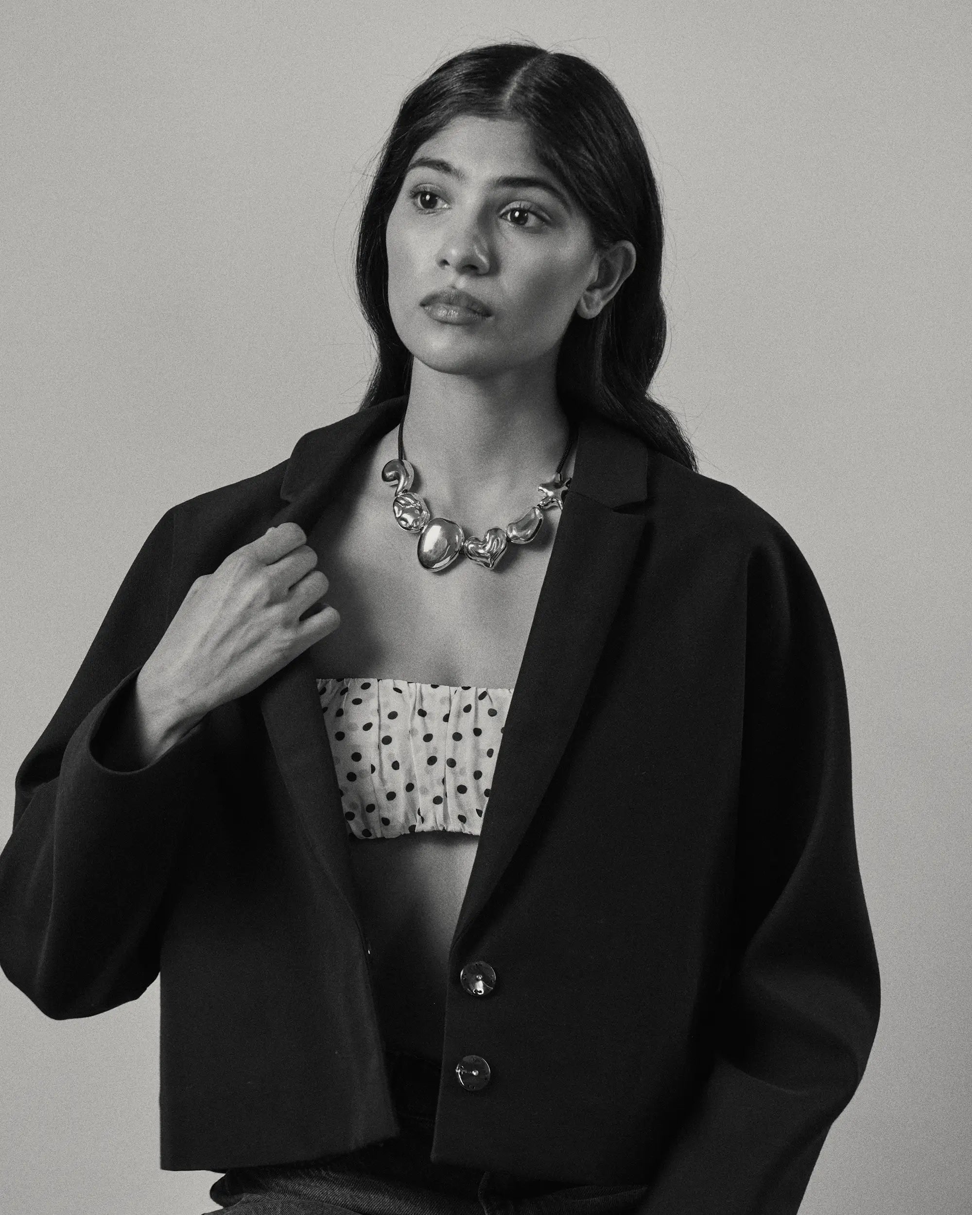 A black and white portrait of a model wearing a black cropped blazer over a white top with black polka dots, accessorized with all 18K Gold Hero objects.