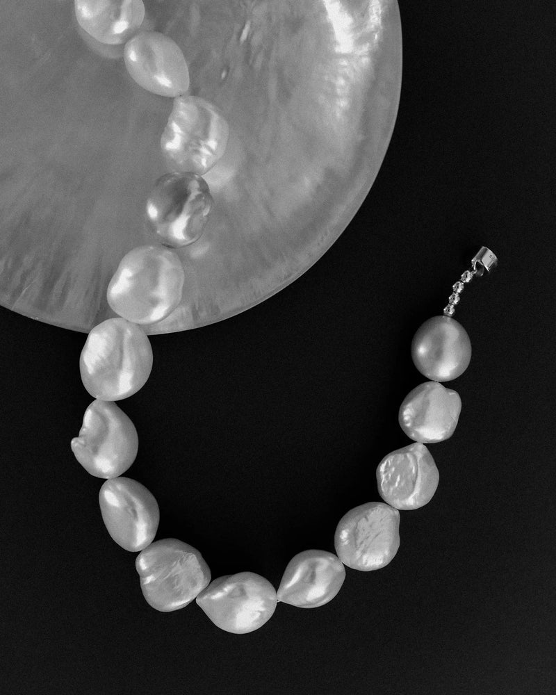 A baroque pearl necklace with irregular, glossy white pearls on a 18K gold clasp, displayed on a black background with a large iridescent shell fragment prop.