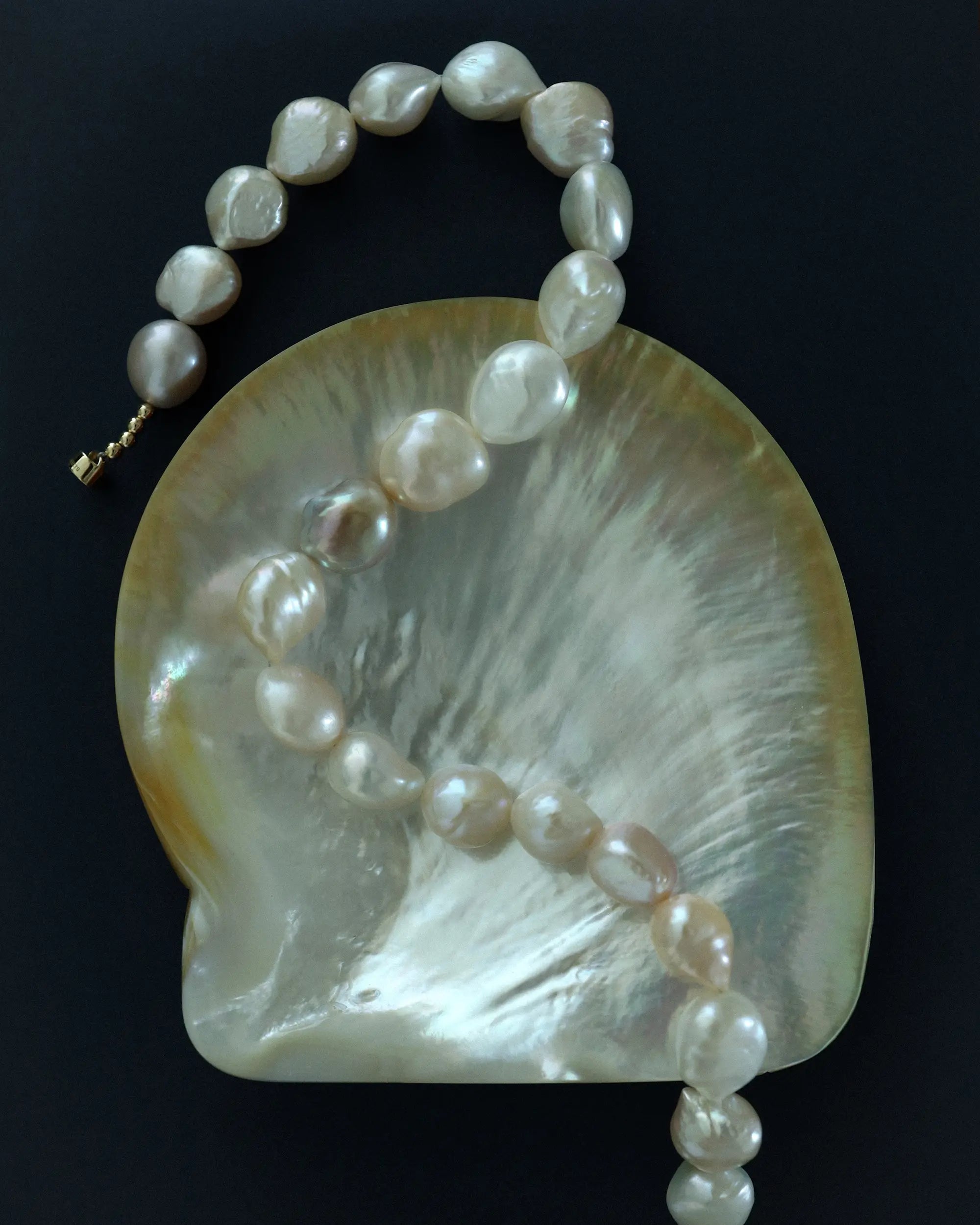 A strand of 18K gold baroque-style freshwater pearls draped over a large iridescent shell, photographed against a solid blue background.