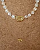 18K Gold Ivory Baroque pearl necklace, 18K Gold Hero Pomegranate pendant and 18K Gold Mini Hero Heart spinner necklace layered on a sand.