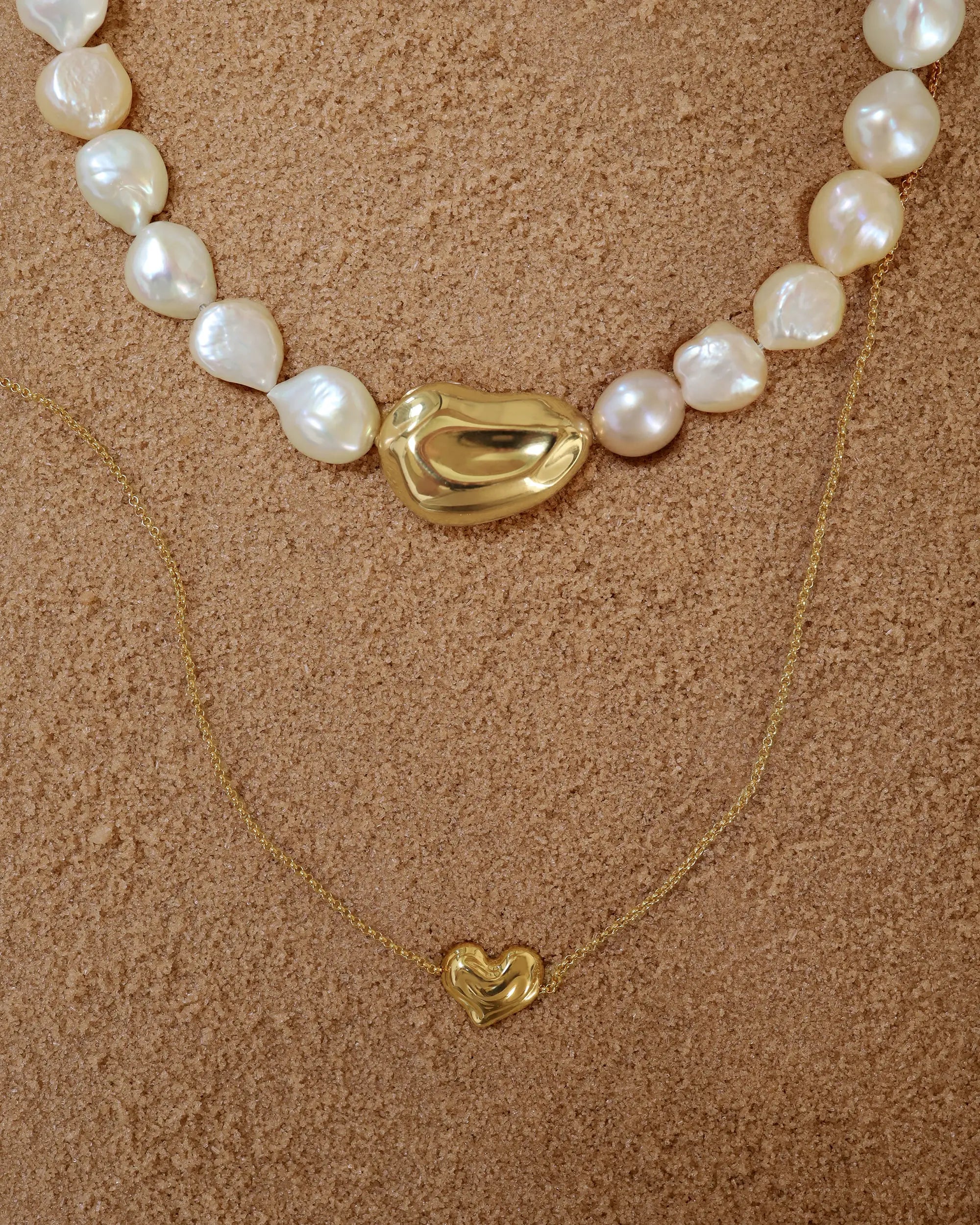 18K Gold Ivory Baroque pearl necklace, 18K Gold Hero Pomegranate pendant and 18K Gold Mini Hero Heart spinner necklace layered on a sand.