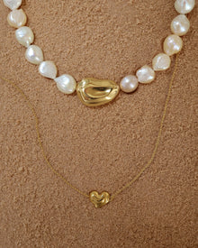 18K Gold Ivory Baroque pearl necklace, 18K Gold Hero Pomegranate pendant and 18K Gold Mini Hero Heart spinner necklace layered on a sand.