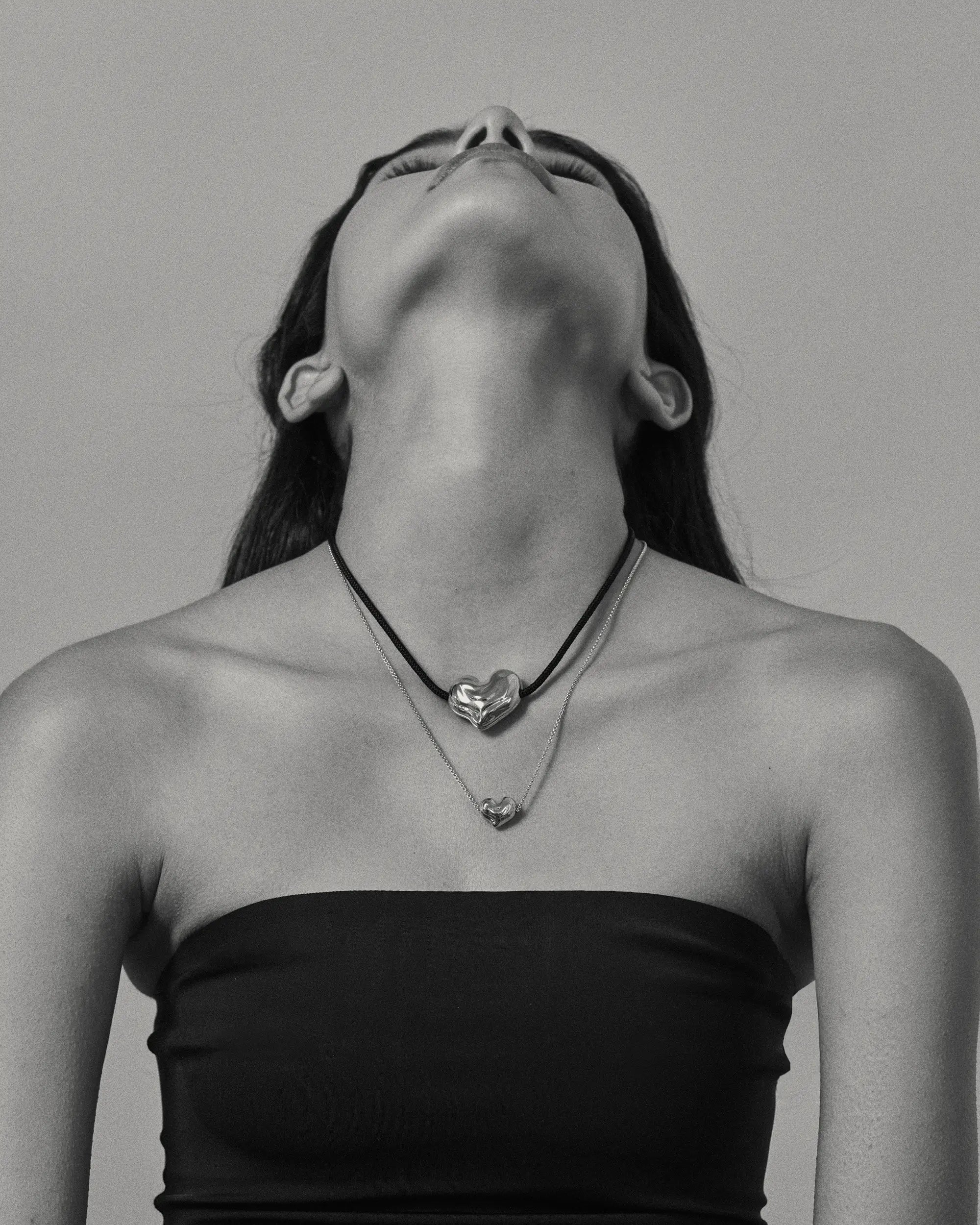 Black and white photo of a model wearing 18K gold black cord necklace with a 18K gold heart-shaped pendant and mini 18K gold chain heart-shaped pendant