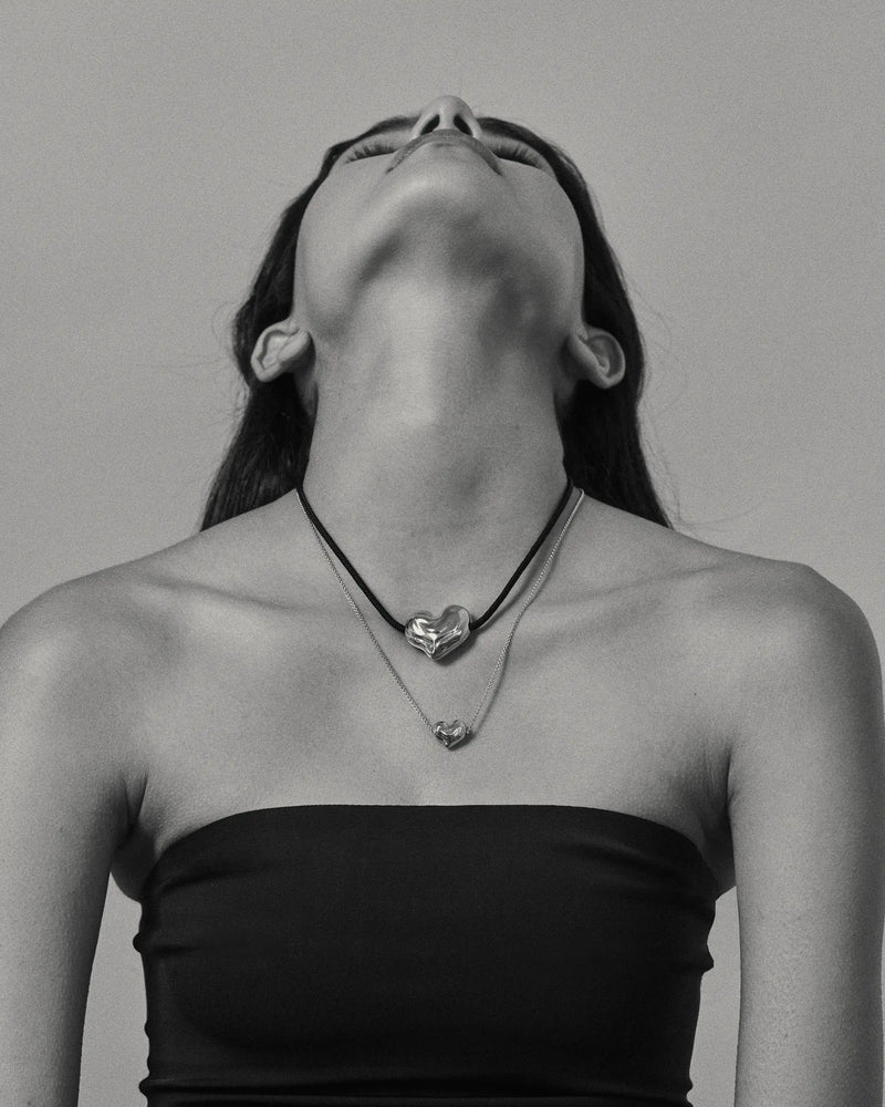 Black and white photo of a model wearing 18K gold black cord necklace with a 18K gold heart-shaped pendant and mini 18K gold chain heart-shaped pendant