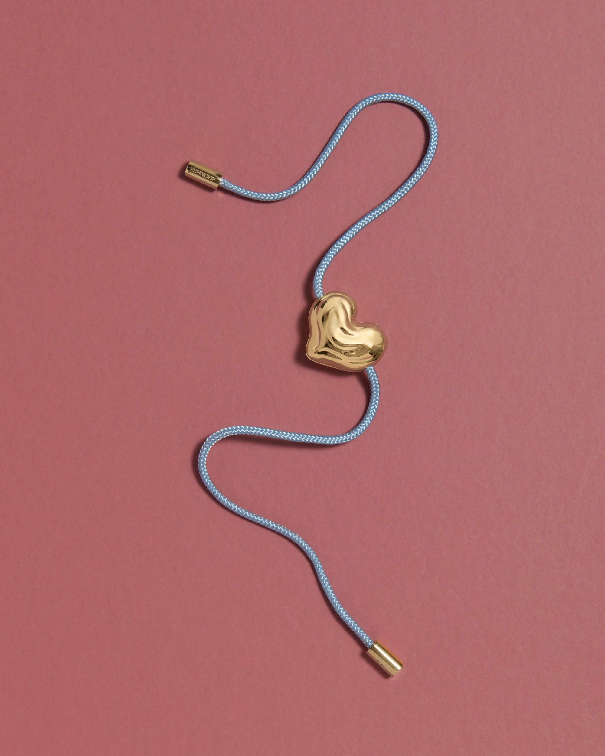 Blue cord necklace with a 18K gold clasp and a heart-shaped 18K gold pendant on a pink background