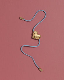 Blue cord necklace with a 18K gold clasp and a heart-shaped 18K gold pendant on a pink background