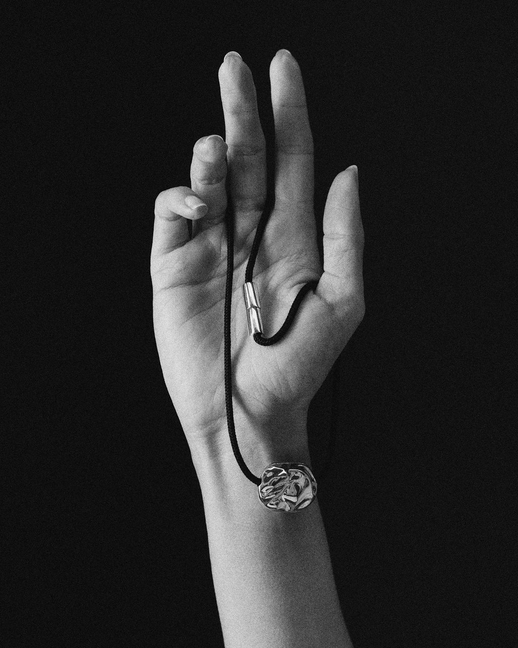 A 18K Gold black cord necklace with a 18K Gold Hero Desert pendant, held in a person's hand against a solid black background.
