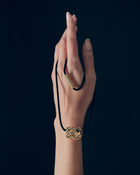 Hand holding desert-shaped 18K gold pendant and 18K gold black cord necklace on a black background