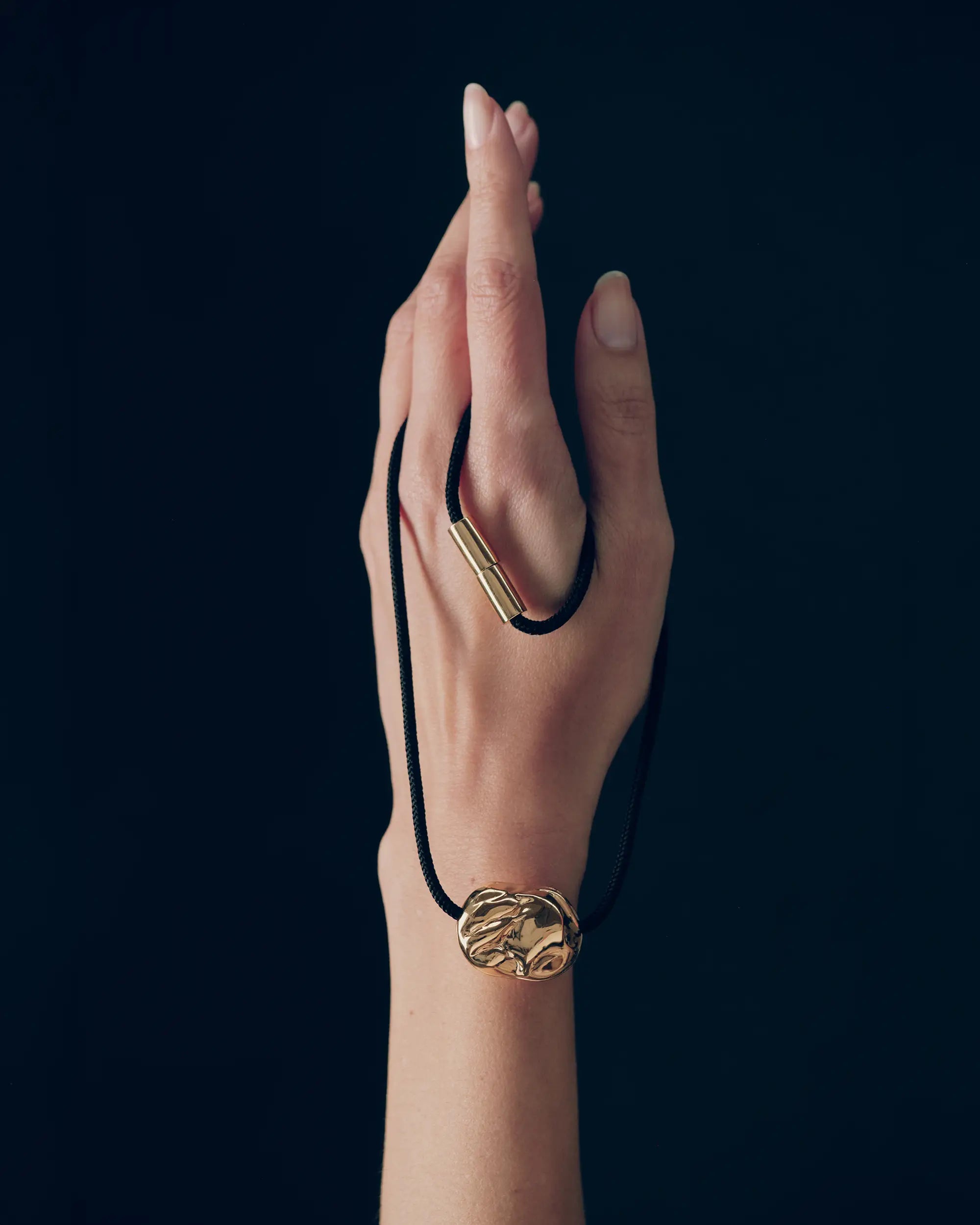 Hand holding desert-shaped 18K gold pendant and 18K gold black cord necklace on a black background