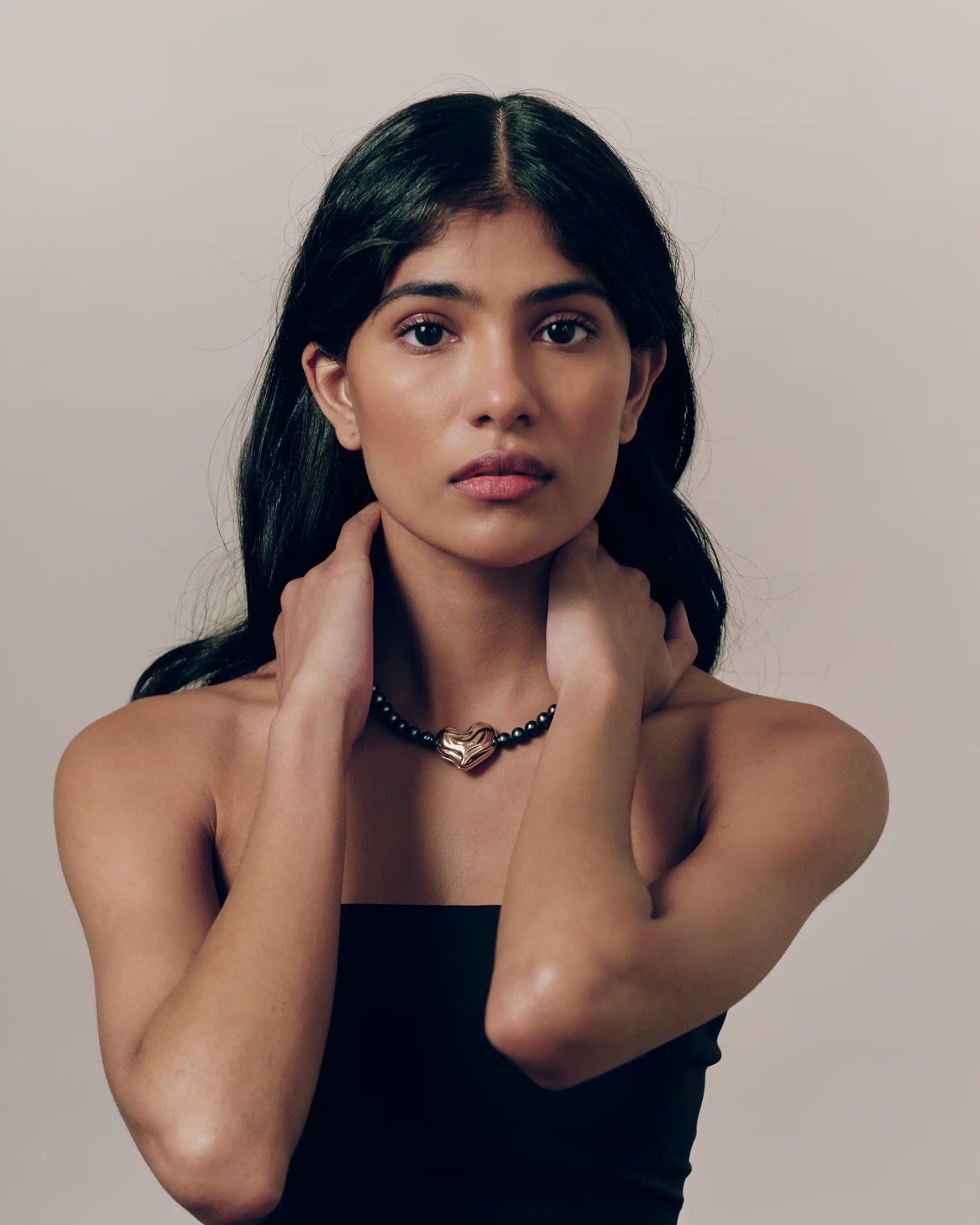 Model wearing 18K gold black pearl necklace and heart-shaped 18K gold pendant on a beige background