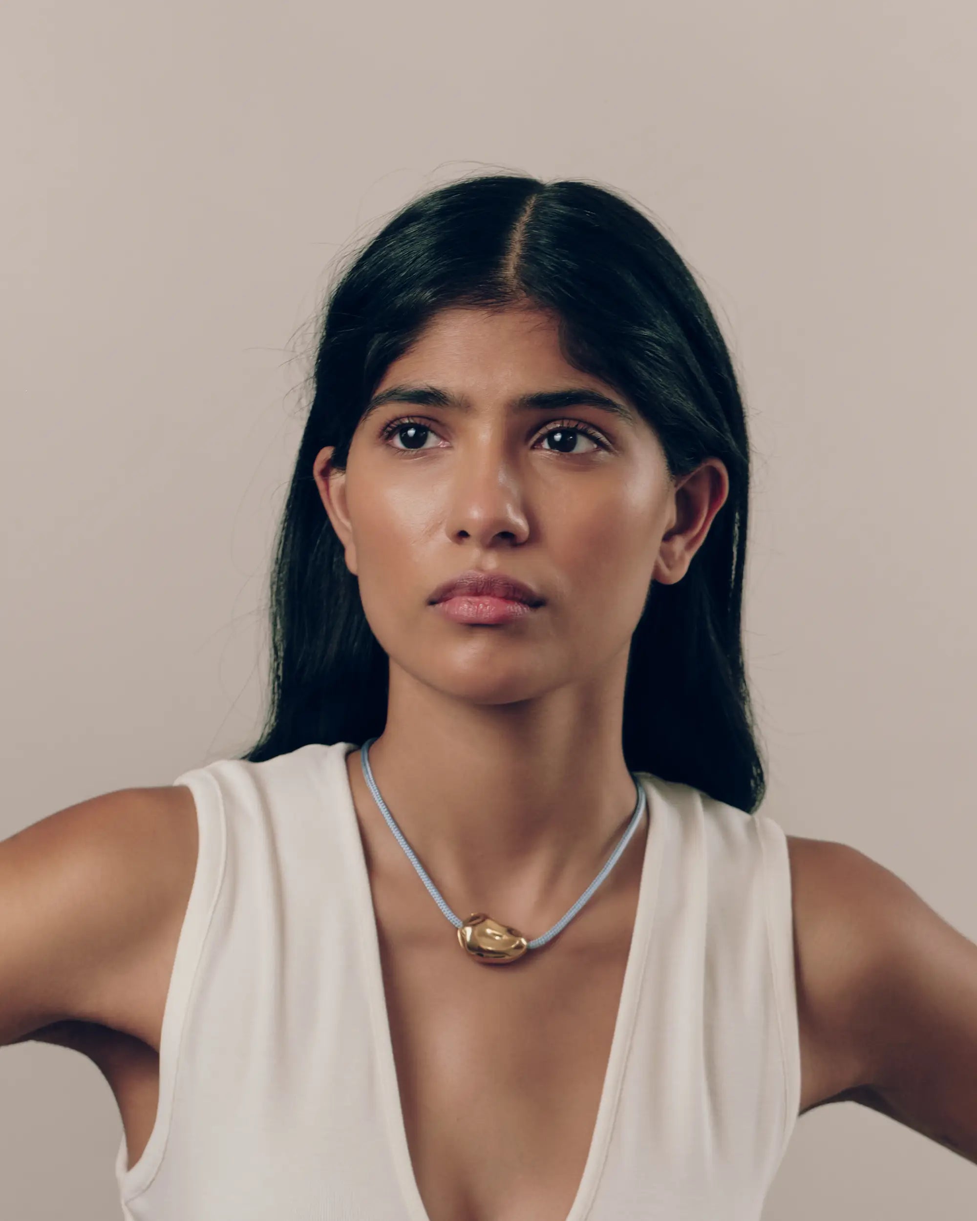 A model with dark hair wearing a white sleeveless top and a 18K gold light blue cord necklace with a 18K gold Hero Pomegranate pendant, photographed against a plain beige background.