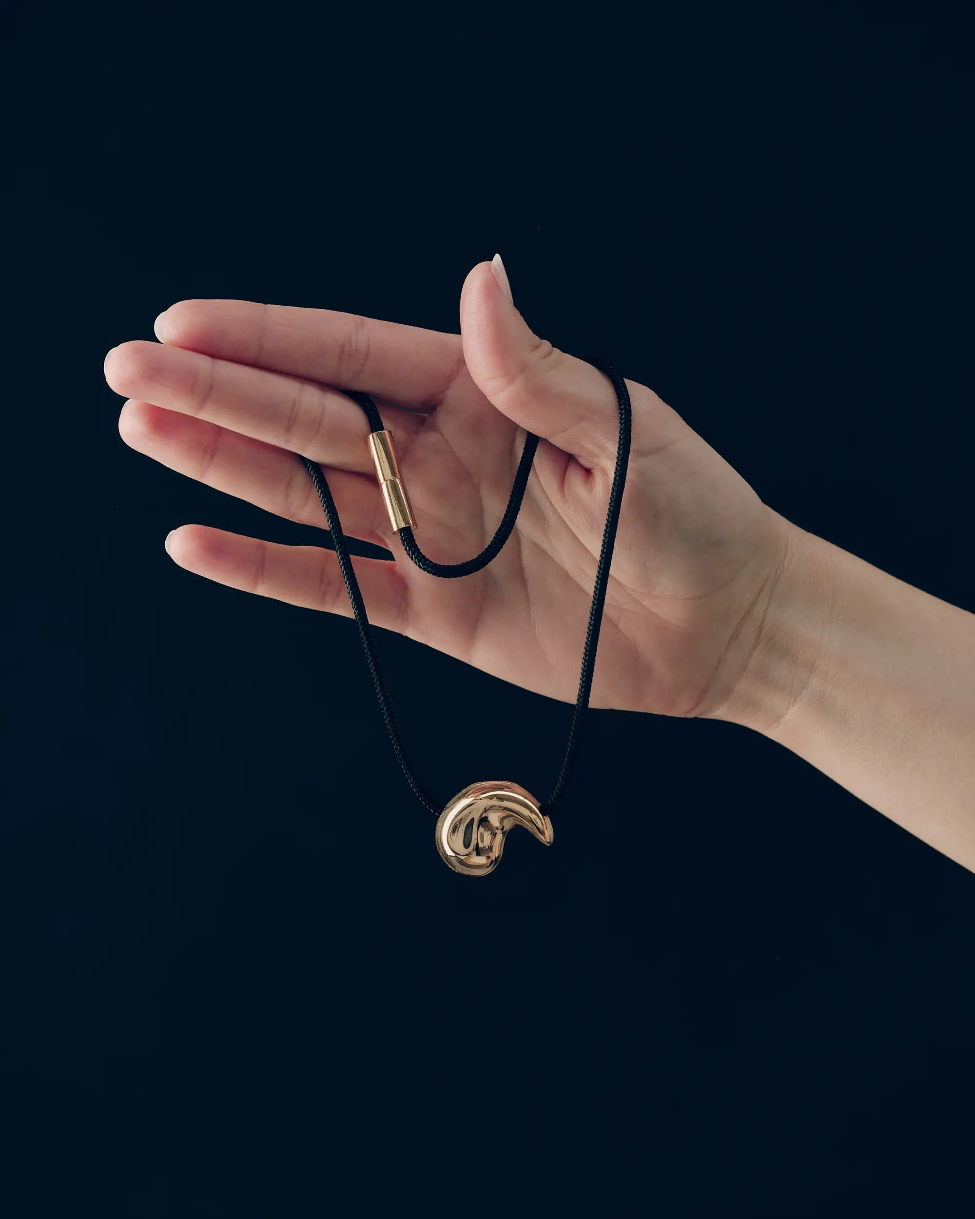 Hand holding spiral-shaped 18K gold pendant and 18K gold black cord necklace on a black background