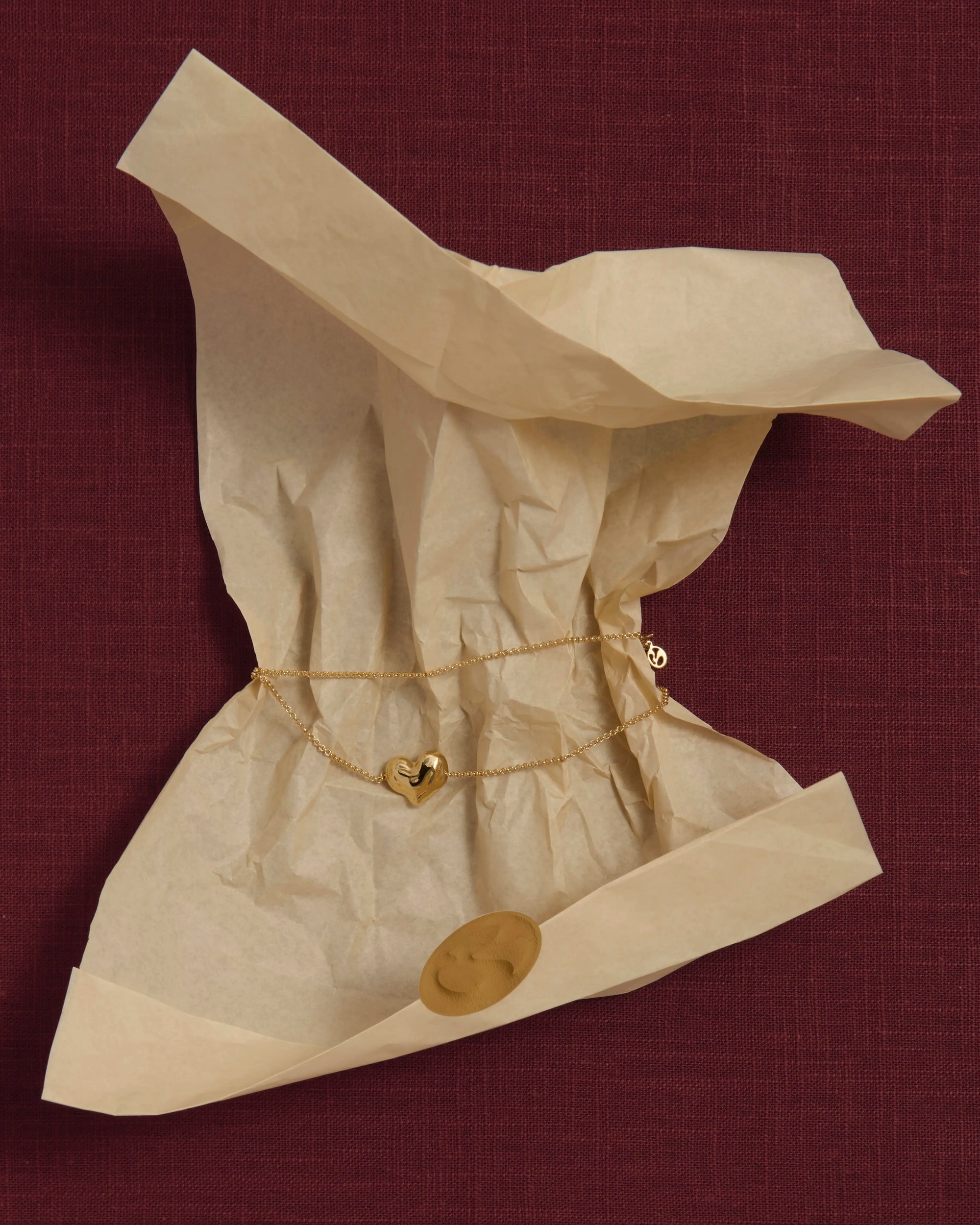 18K gold rollo chain necklace with a heart-shaped pendant on crumpled beige tissue paper against a pink linen background