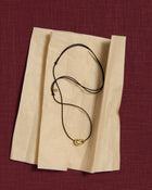 Black cord necklace with a 18K gold pomegranate-shaped pendant on a tissue paper and pink linen background
