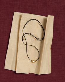 Black cord necklace with a 18K gold pomegranate-shaped pendant on a tissue paper and pink linen background