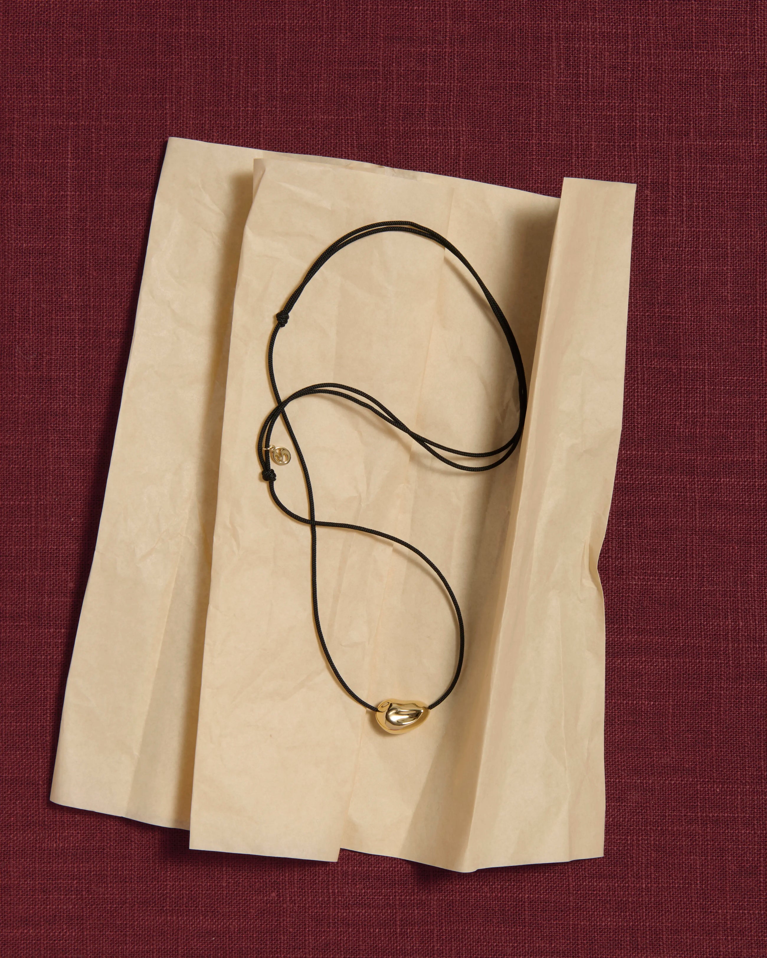 Black cord necklace with a 18K gold pomegranate-shaped pendant on a tissue paper and pink linen background