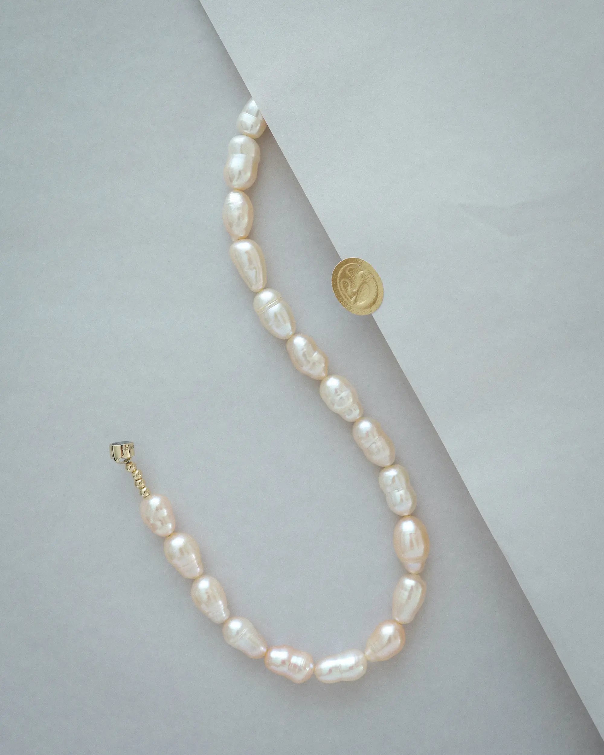 A 18K gold peach pearl necklace with irregular, teardrop-shaped pearls, displayed on a light grey background with a gold sticker prop.