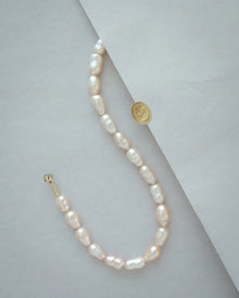 A 18K gold peach pearl necklace with irregular, teardrop-shaped pearls, displayed on a light grey background with a gold sticker prop.