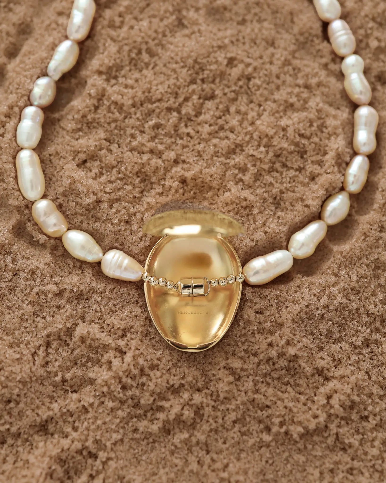 Opened 18K Gold Hero Pebble pendant and 18K Gold Freshwater pearl in Peach on a sand