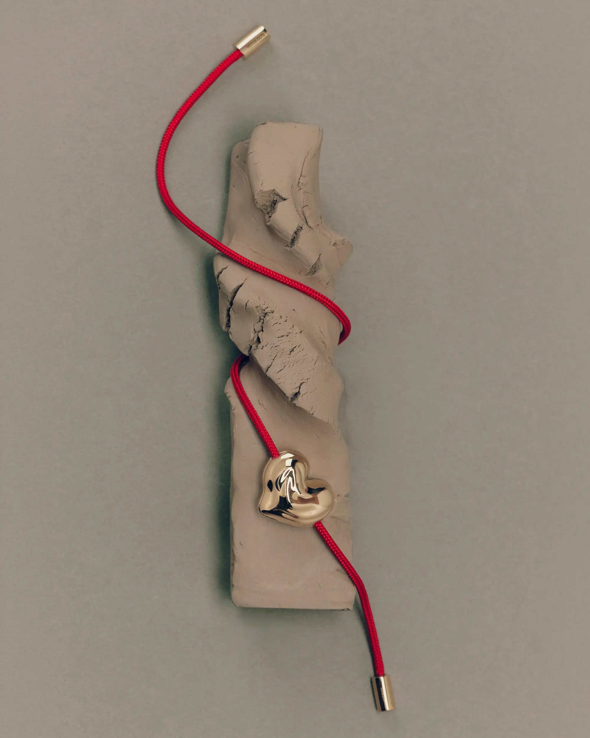 Red cord necklace with a 18K gold clasp and 18K gold heart-shaped pendant draped around clay sculpture