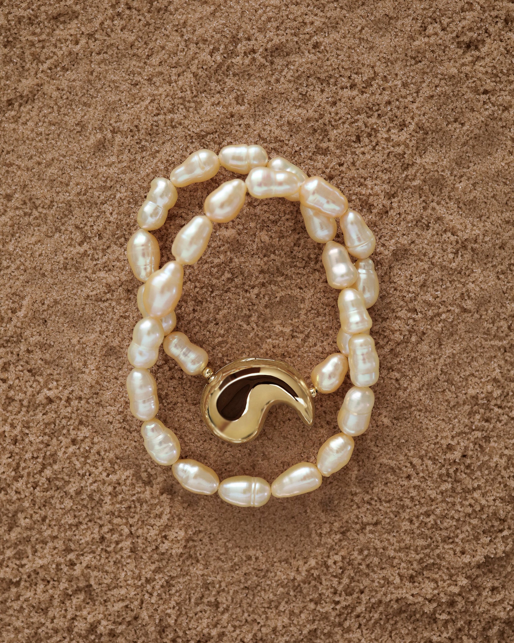 18K Gold Hero Spiral pendant and 18K Gold Peach freshwater pearl on a sand