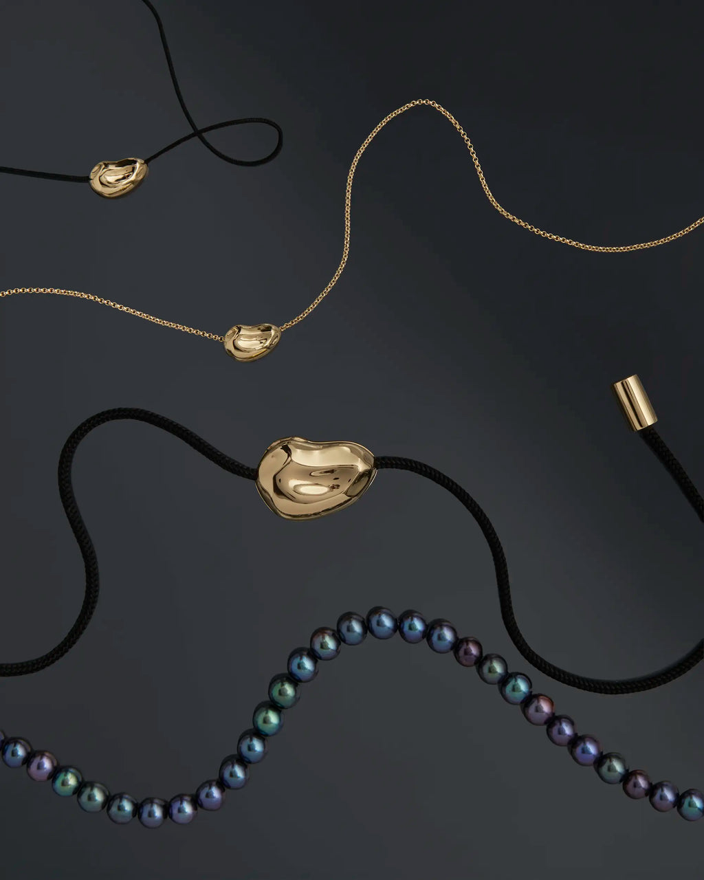 18K gold black cord, 18k gold rollo chain, and 18K gold black pearl necklaces with a 18K gold pomegranate seed pendants agains black background.